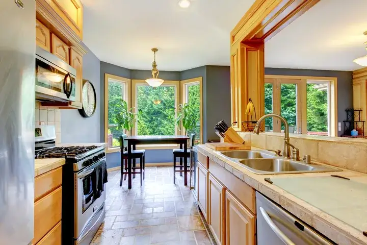 Bright, spacious kitchen with light wood cabinetry, stainless steel appliances, a double sink, and large windows overlooking lush green trees. A small dining table sits in a bay window nook, filling the space with natural light.